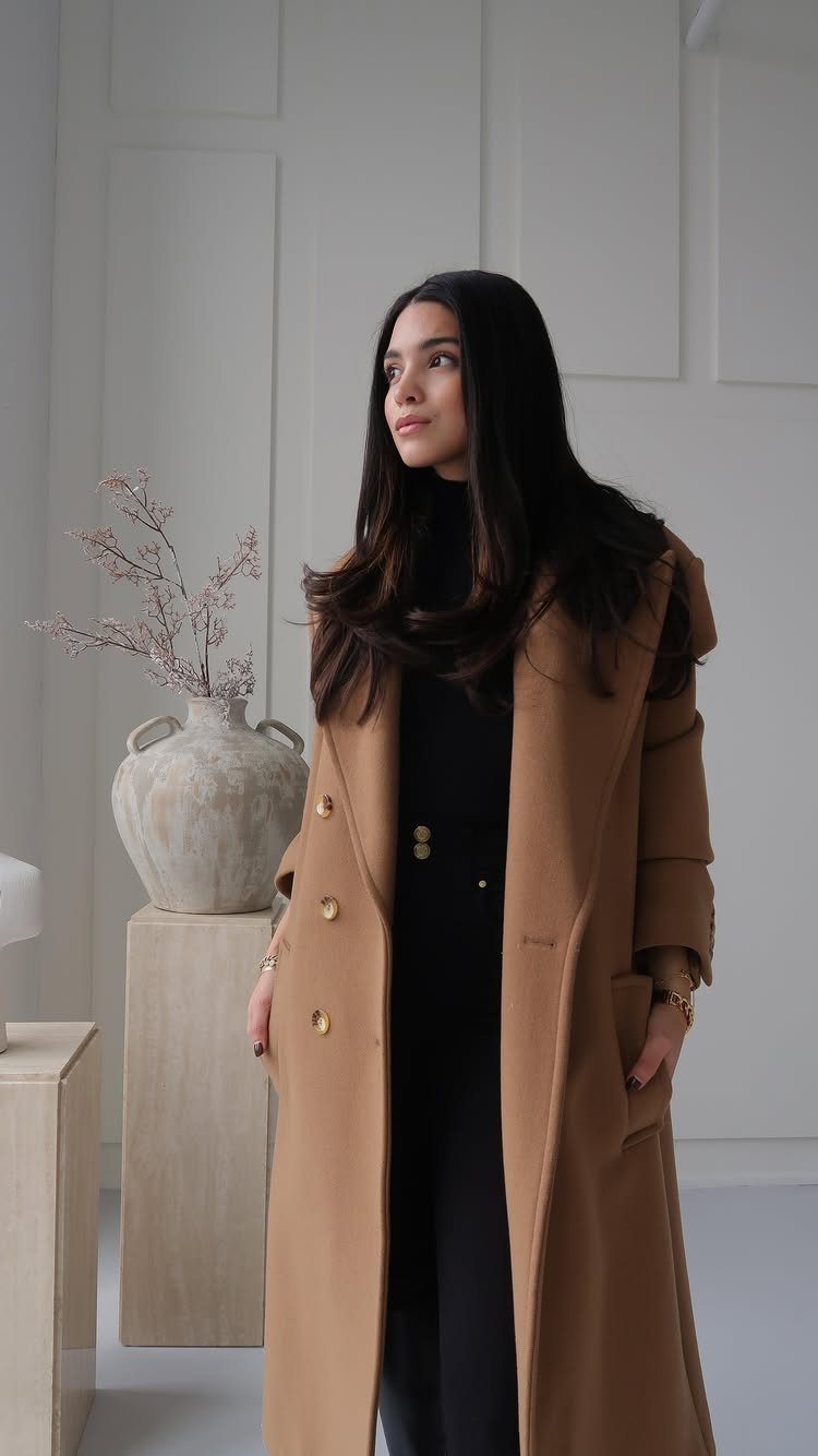 A woman wearing a long tan wool coat and black turtleneck in a minimalist room.