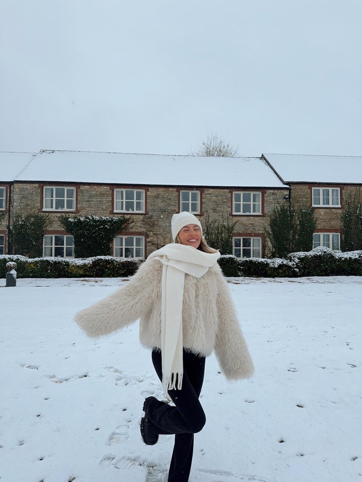 Woman wearing a cream faux fur coat and white scarf posing in a snowy outdoor setting.