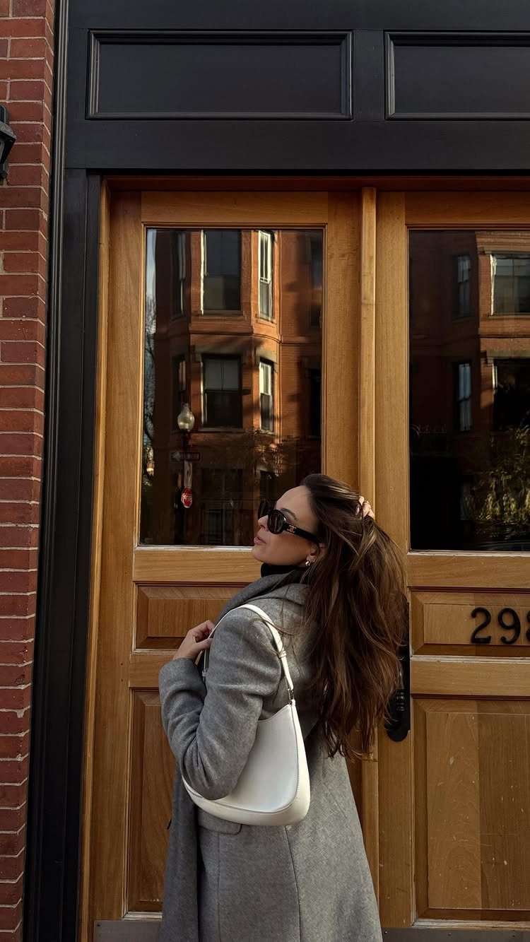Woman in a grey wool coat and sunglasses standing before a wooden door.