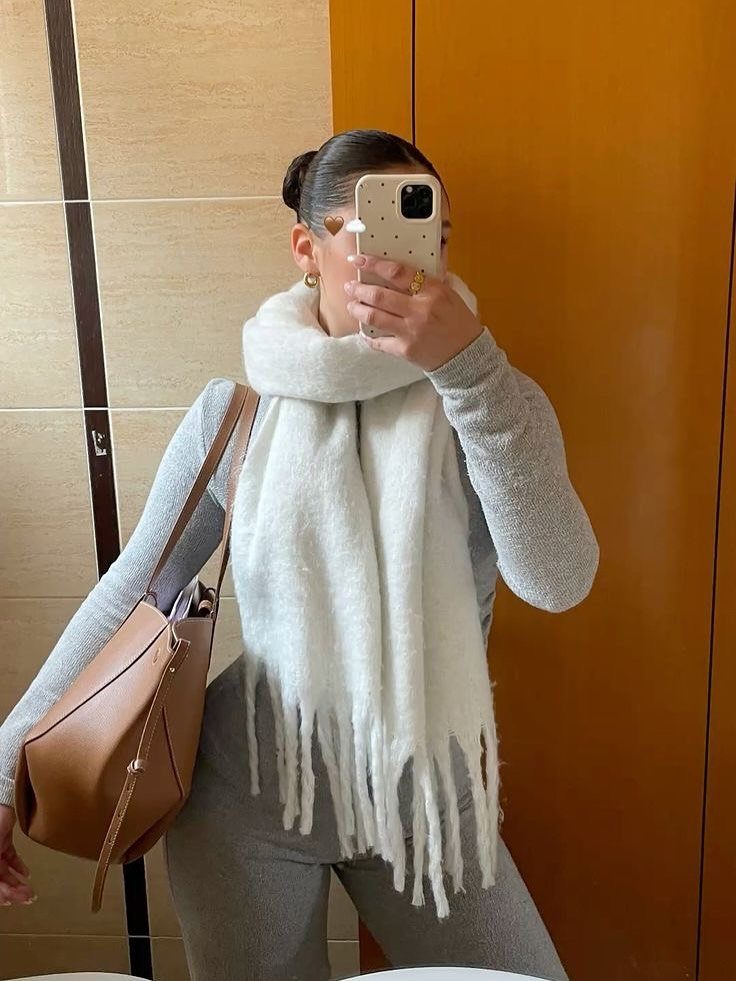Woman in grey outfit wearing a large white fringed scarf and carrying a tan handbag.