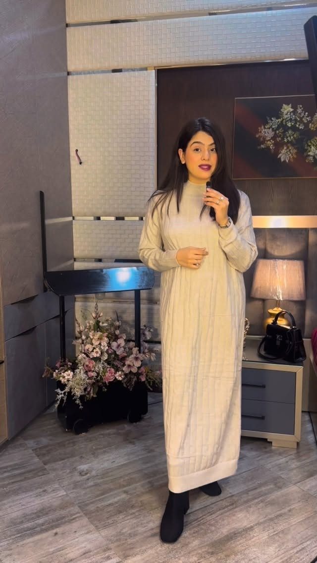 A woman posing in a long cream ribbed sweater dress and black boots indoors.