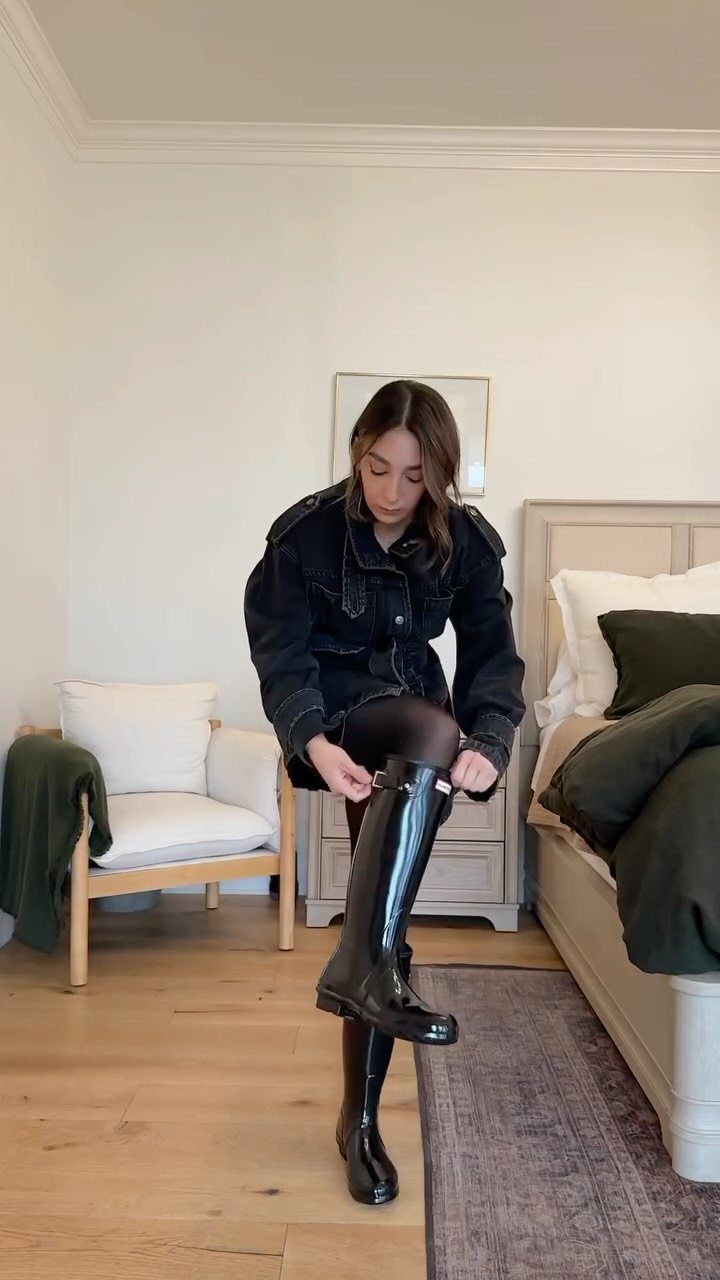 A woman putting on tall, shiny black waterproof boots while wearing a black denim jacket.