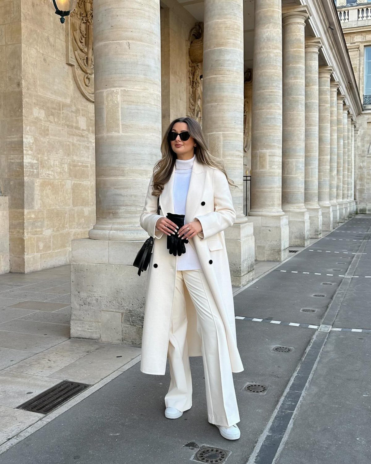A woman posing in a long white coat, white turtleneck, and white trousers against stone columns.
