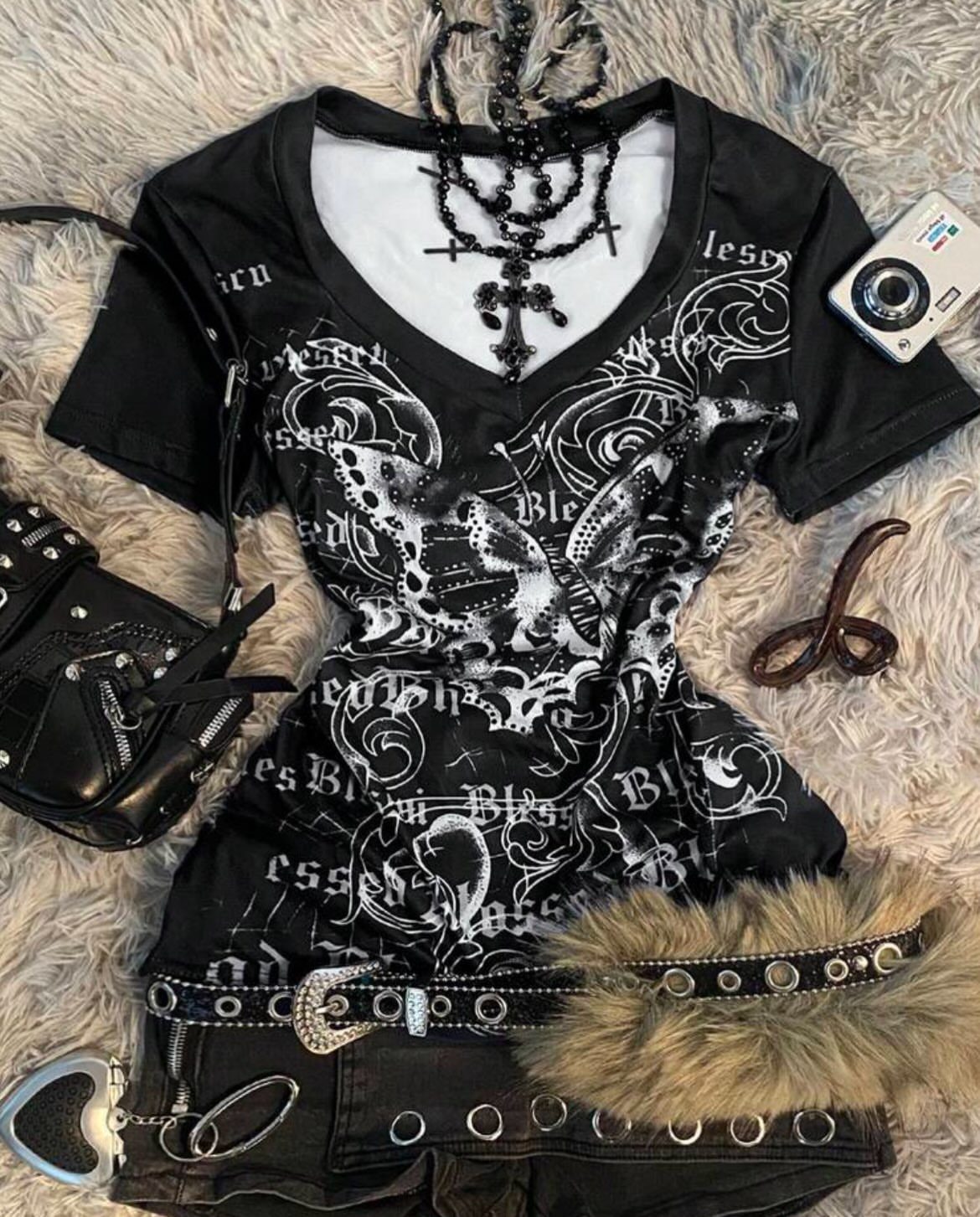 Black graphic t-shirt, studded belt, denim shorts, and accessories arranged on white faux fur.