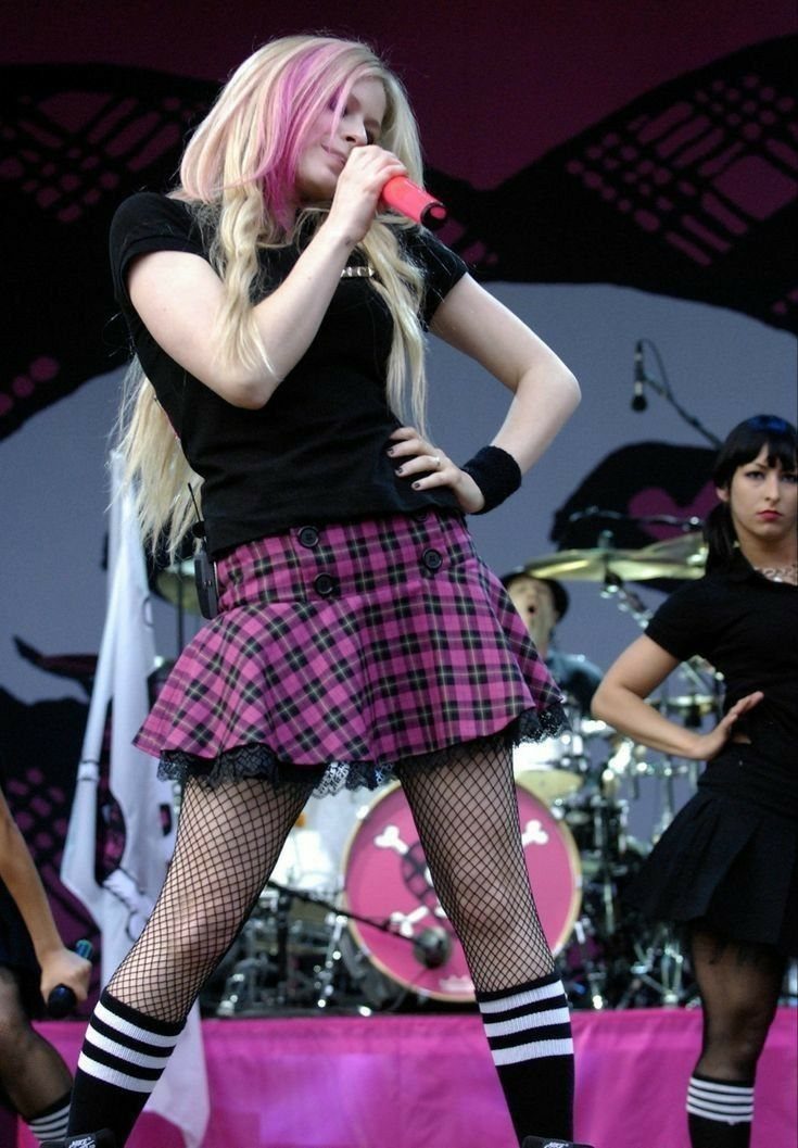 A singer wearing a black t-shirt and pink plaid pleated mini skirt performs on stage.