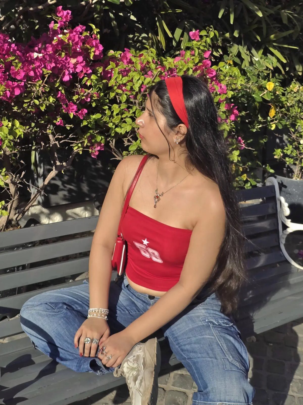 A woman in a red slogan tube top and baggy blue jeans sitting on a bench.
